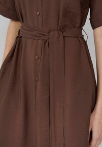 Hněd dress s texturou a krátkými rukávy, s uvázaným páskem v pase, zobrazený od hrudi po boky, nošený osobou s uvolněnými pažemi po stranách.