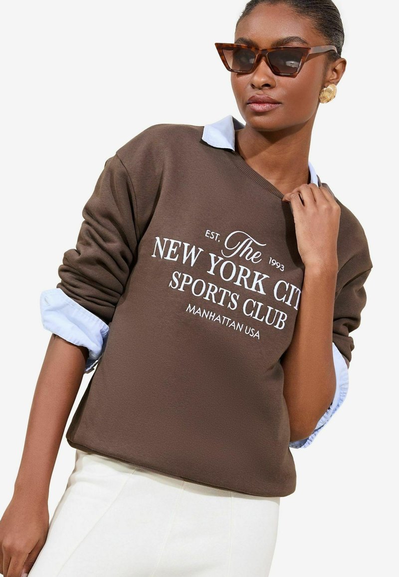 Felpa marrone con testo bianco che legge "The New York City Sports Club" sopra una camicia con colletto azzurro chiaro, con polsini e orlo a coste.