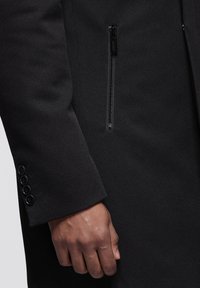 Zwarte textuur jas met een ritssluiting zak op de borst en knoopmanchetten, met een gladde stofafwerking en een getailleerd ontwerp.