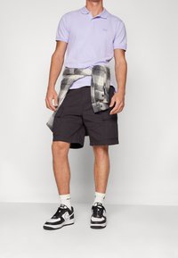 Polo violet clair avec logo GAP, chemise à carreaux grise nouée autour de la taille, shorts cargo foncés, baskets noires et blanches, chaussettes blanches.