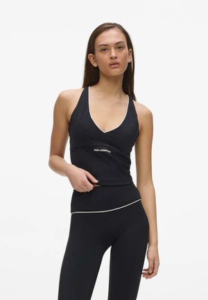 Kvinde iført sort ærmeløs top med V-hals og sorte leggings med hvide detaljer, stående mod en ensfarvet hvid baggrund.