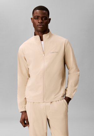 Homme portant une veste beige zippée et un pantalon assorti, debout devant un fond gris uni.