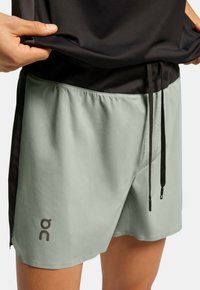 Pantalones cortos de color verde claro con una textura suave, cintura elástica y cordón ajustable. Presentan un panel lateral negro y el logo "on".