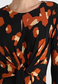 Top noir avec un détail en torsion à l'avant, présentant un imprimé léopard abstrait orange et crème, encolure ronde et accent noué à la taille.