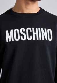Sudadera negra con el distintivo texto blanco "MOSCHINO" en el pecho, hecha de un material de algodón suave y con un diseño de cuello redondo.