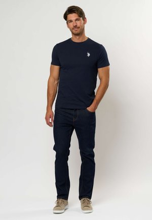 Mørkeblå bomulds T-shirt med rund hals og hvidt logo, kombineret med mørke denimjeans og lyse beige sneakers. Enkel, tætsiddende pasform.