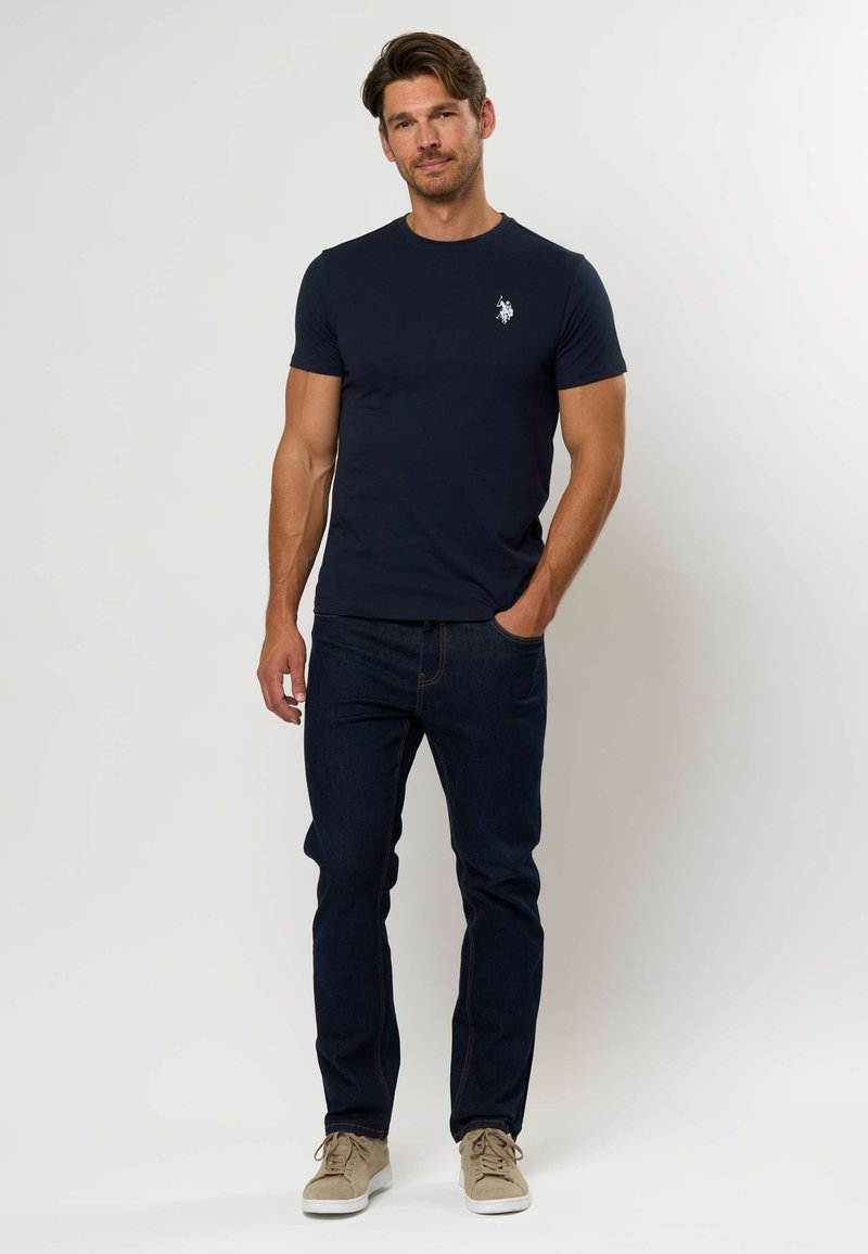 Tamsiai mėlynas medvilnės marškinėliai su apvalia iškirpte ir baltu logotipu, derinami su tamsiais džinsais ir šviesiai smėlio spalvos sportbačiais. Paprastas, prigludęs dizainas.