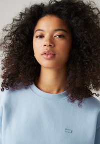 Sweatshirt bleu clair avec un col rond, fabriqué en tissu doux. Présente un petit logo Levi's brodé sur la poitrine. Cheveux bouclés au premier plan.