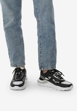 Sneaker atletiche in bianco e nero con accenti grigi, caratterizzate da una parte superiore testurizzata, chiusura con lacci e suola spessa bianca. Indossate con jeans azzurri chiari.