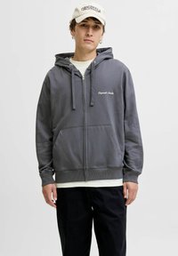 Sudadera gris con cremallera, bolsillos canguro, capucha con cordones y puños acanalados. Lleva puesta sobre una camiseta crema y cuenta con un logo bordado en el pecho.