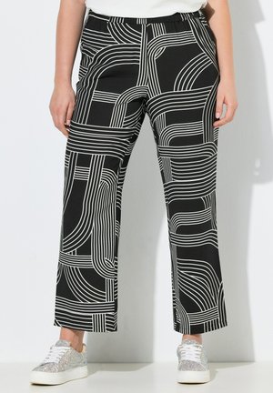 Zwarte broek met opvallende witte geometrische lijnenpatronen, gedragen met zilverkleurige glinsterende sneakers en een wit shirt.