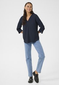 Blusa blu navy a V con texture e maniche tre quarti, abbinata a jeans azzurri e scarpe con plateau nere, in piedi su uno sfondo bianco.