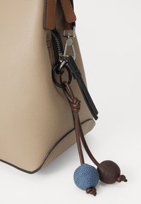 Borsa a mano in pelle beige con superficie testurizzata, dotata di una cerniera, manici marroni e neri, e pompon decorativi blu e marroni.