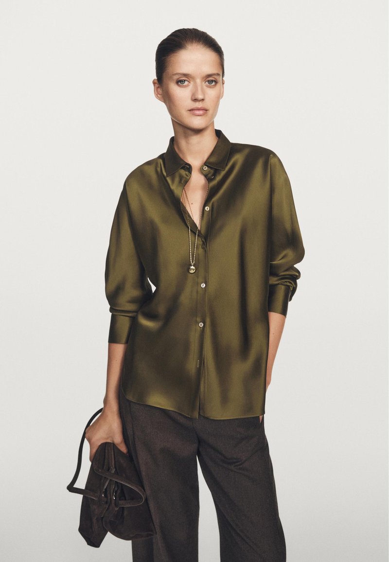 Olive green satin blouse with a collar, button-down front, and long sleeves, paired with dark trousers and a brown bag.