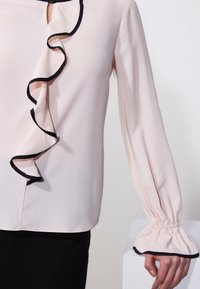 Blusa rosa chiaro con rifiniture in nero arricciate lungo il collo e le maniche a polsino. Presenta un'apertura a chiave sul collo. Tessuto dalla superficie liscia.