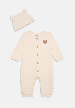 Cream-colored baby romper with long sleeves, ribbed texture, brown bear patch, wooden buttons, and matching hat with bear ears.