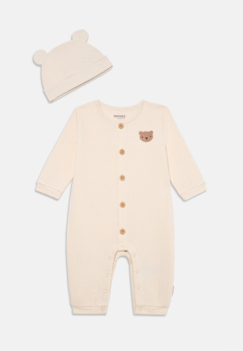Salopetă pentru bebeluși de culoare crem, cu mâneci lungi, textură ribbed, patch cu ursuleț maro, nasturi din lemn și căciulă asortată cu urechi de urs.