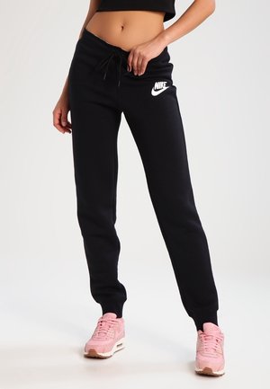 Person wearing black Nike sweatpants and pink Nike sneakers, standing with one hand adjusting the waistband.