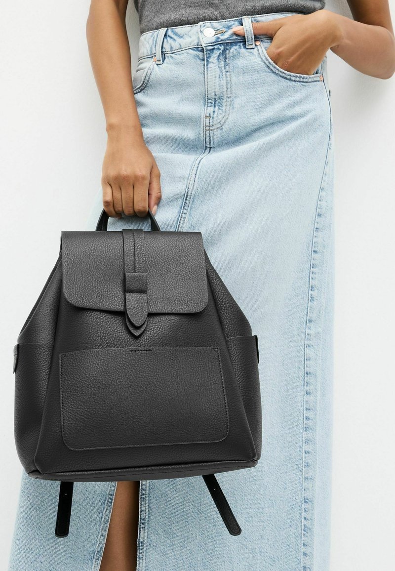 Femme en jupe en jean bleu clair tenant un sac à dos en cuir noir par sa poignée supérieure sur un fond uni.