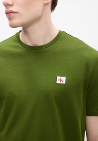 Zöld pamut póló kerek nyakkivágással, bal oldalon egy kis fehér logóval és piros felirattal. Simaság jellemzi, laza kialakítás.