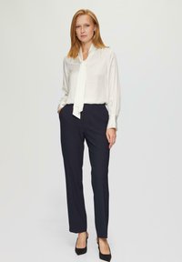 Blouse blanche avec un nœud, manches longues ; associée à un pantalon bleu marine et des talons noirs. Tissu lisse, coupe ajustée, design minimaliste.