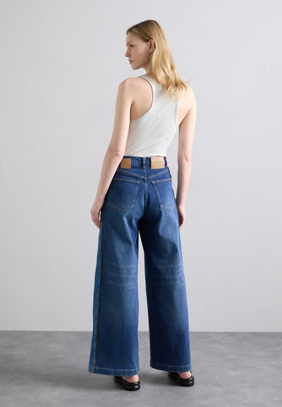 Hellblaues Tanktop mit weit geschnittenen blauen Jeans. Die Jeans haben eine hohe Taille, zwei Gesäßtaschen und ein strukturiertes braunes Etikett am Bund.