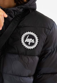 Czarna kurtka puchowa z okrągłą, haftowaną naszywką z napisem "hype" umieszczoną w wieńcu laurowym, teksturowanym materiałem i widocznymi suwakami.