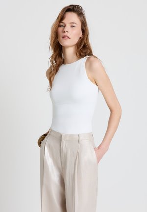 Donna con capelli castani lunghi che indossa una canottiera bianca senza maniche e pantaloni beige lucidi a vita alta, con le mani nelle tasche.