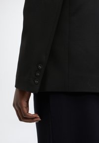 Manica di blazer nero con tre bottoni rivestiti in tessuto, texture liscia, design strutturato, su sfondo scuro di pantaloni attillati.