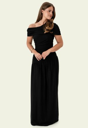 Femme aux longs cheveux ondulés portant un haut noir ajusté, épaules dénudées, et une longue jupe, debout, les mains jointes, regardant sur le côté.
