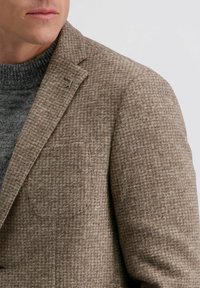 Braun karierter Blazer mit einem strukturierten Stoff, ausgestattet mit einer Brusttasche und einem silbernen Akzentdetail am Revers, kombiniert mit einem grauen Pullover.