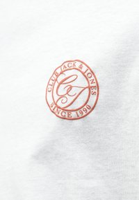 Camisa blanca de manga corta con un logo bordado circular en naranja que dice "CLUB JACK & JONES" y "DESDE 1990." Textura de tejido suave.