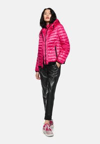 Woman standing with hands in pockets, wearing bright pink quilted jacket, black leather-look pants, and pink sneakers against a white background.