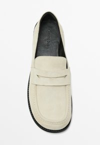 Light beige suede loafers with a rounded toe, stitch detailing, and a decorative strap. Black interior with logo. Flat rubber sole.