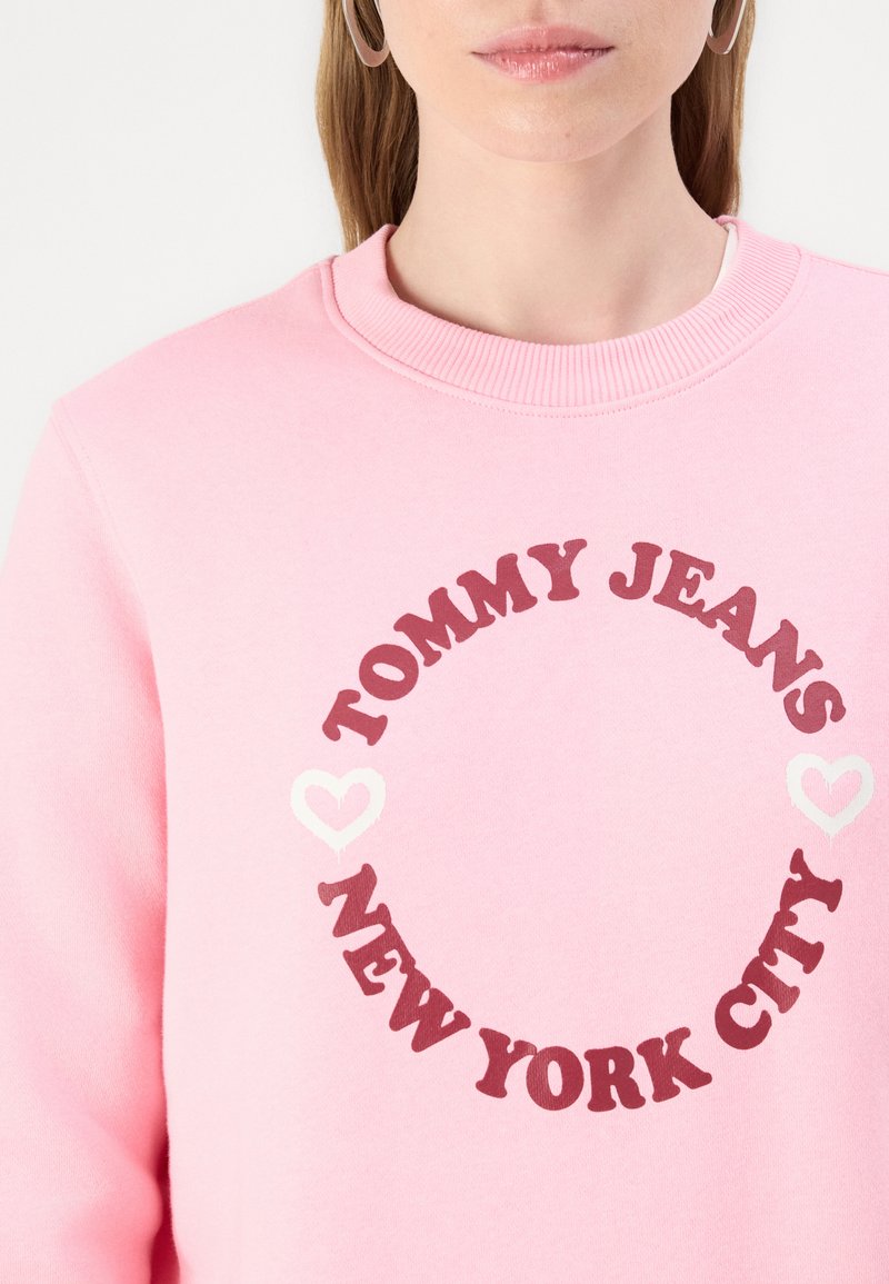 Felpa rosa con colletto a coste, caratterizzata dalla scritta "TOMMY JEANS" e "NEW YORK CITY" in rosso scuro, contornata da due cuori bianche.