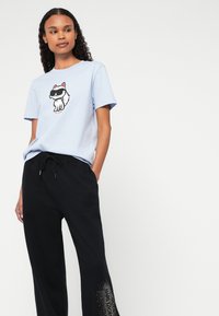 Ανοιχτό μπλε βαμβακερό t-shirt με γραφικό μαύρης και λευκής γάτας, συνδυασμένο με μαύρα παντελόνια που διαθέτουν κορδόνι και διακοσμητικά στοιχεία στο τελείωμα.