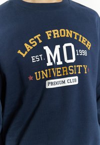 Felpa blu navy in cotone, con colletto a girocollo. La parte frontale mostra del testo bianco, giallo e rosso, comprensivo di "LAST FRONTIER UNIVERSITY."