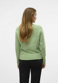 Maglione verde lavorato a maglia con una texture morbida, design aderente e polsini a coste. Indossato con pantaloni di velluto a coste neri, visto da dietro.
