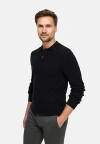 Schwarzes langärmliges Poloshirt mit strukturiertem Strickmuster, ausgestattet mit einem Kragen und einer Knopfleiste mit zwei Knöpfen, kombiniert mit grauen maßgeschneiderten Hosen.