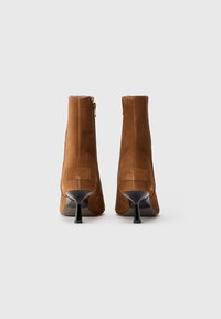 Stivaletti alla caviglia in pelle scamosciata marrone con cerniera laterale, punta affilata e tacco a stiletto nero. Presentano una texture liscia e un minimo dettaglio di branding.