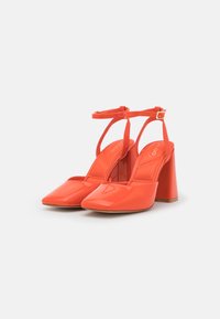 Sandales à talons hauts en cuir verni corail avec un nez pointu, une bride de cheville et un talon épais. Texture lisse avec des accents minimaux.