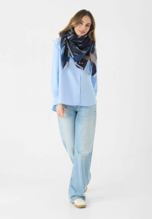 Chemise à manches longues bleu clair avec des boutons, associée à un jean en denim clair ample. Une écharpe à motifs bleu marine et beige drape autour du cou.