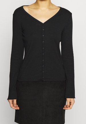 Femme portant un haut noir cintré à manches longues avec boutons, col en V, et une jupe noire sur un fond uni clair.