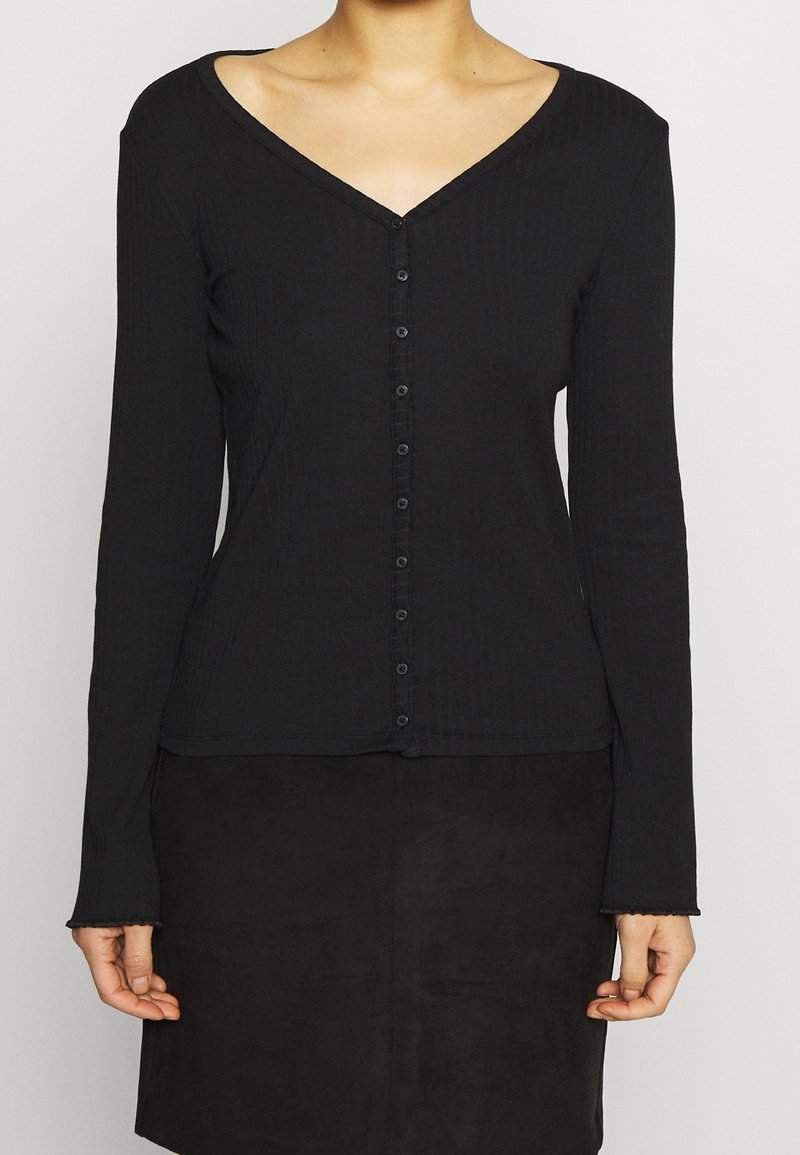 Femme portant un haut noir cintré à manches longues avec boutons, col en V, et une jupe noire sur un fond uni clair.