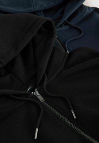 Dos sudaderas: una negra y otra azul marino. Ambas cuentan con cordones, cremalleras y una textura suave. Están apiladas de cerca, mostrando sus capuchas y cremalleras.