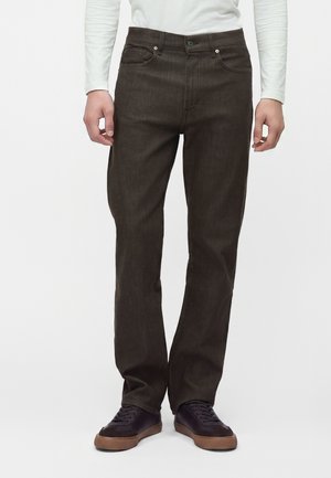Homme portant un jean droit marron foncé avec des baskets noires et marron, debout devant un fond blanc uni.