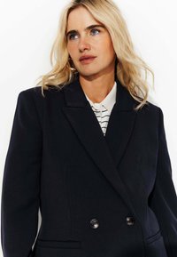 Femme blonde aux yeux bleus portant un blazer noir à double boutonnage par-dessus une chemise rayée blanche et noire, regardant légèrement sur le côté.