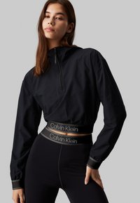 Czarna krótka bluza z suwakiem z przodu na ćwierć długości i teksturowanym materiałem. Połączona z czarnymi legginsami o wysokim stanie z logo w pasie.