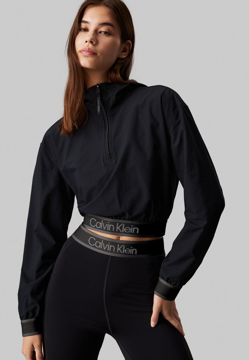 Czarna krótka bluza z suwakiem z przodu na ćwierć długości i teksturowanym materiałem. Połączona z czarnymi legginsami o wysokim stanie z logo w pasie.