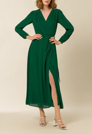 Frau trägt ein langärmeliges grünes Wickelkleid mit einem Oberschenkel hohen Schlitz und weißen offenen Blockabsatz-Sandalen, steht vor einem neutralen Hintergrund.